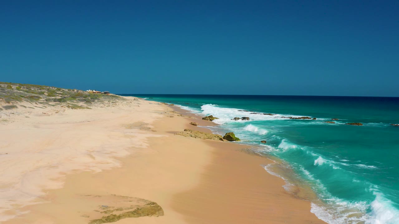 Aerial view at El Cardoncito with turquoise water filling more of the right frame, foamy surf reaching the sand, scrub-covered dunes in the distance, and no visible people.