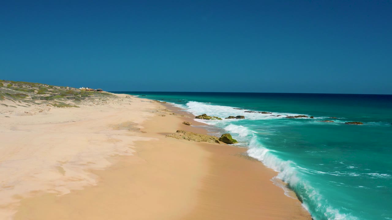 Aerial frame at El Cardoncito with a broad empty beach, white surf curving along the waterline, rocks near the midground, and no visible people.