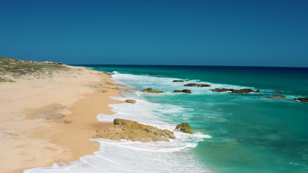 Aerial view over El Cardoncito beach with tan sand on the left, turquoise waves breaking around rocks, a clear horizon, and no visible people.