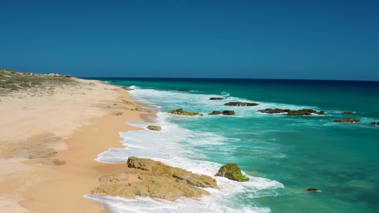 Aerial frame over El Cardoncito showing a wider section of turquoise water, white foam spreading across the beach edge, dark rocks offshore, and no visible people.