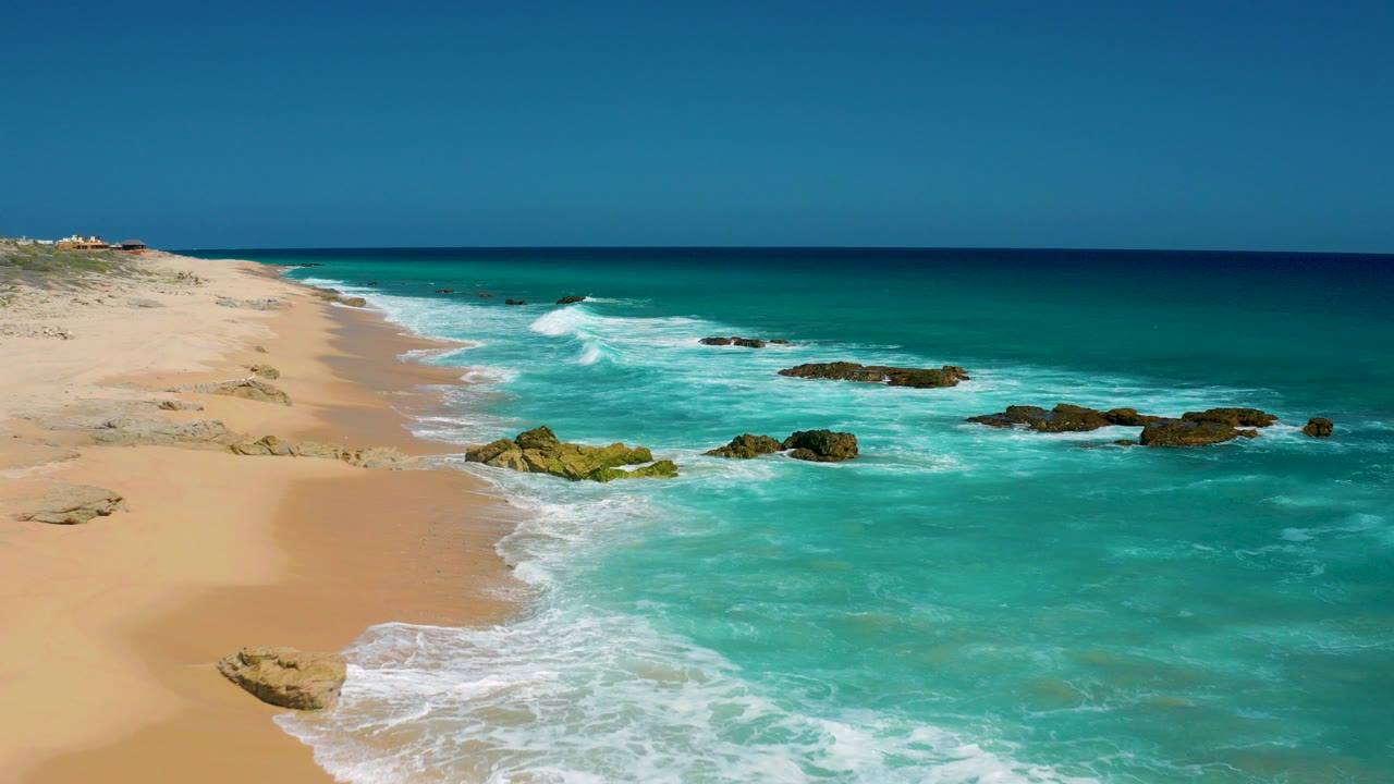 Opening frame of El Cardoncito, Baja California Sur beach with empty sand on the left, turquoise water on the right, scattered rocks in the surf, and no visible people.