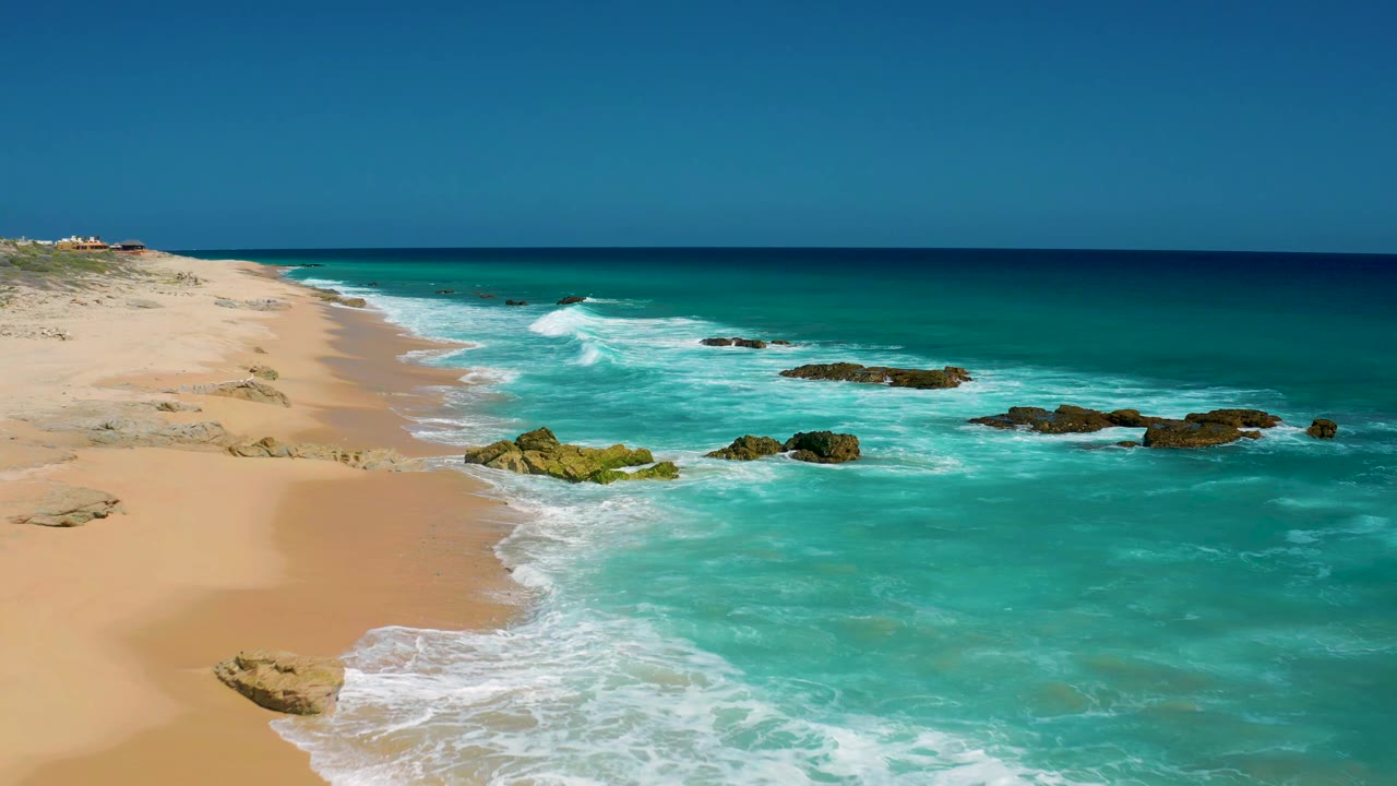 El Cardoncito shoreline continues with pale sand curving into turquoise surf, low rocks near the waterline, a clear horizon, and no visible people.