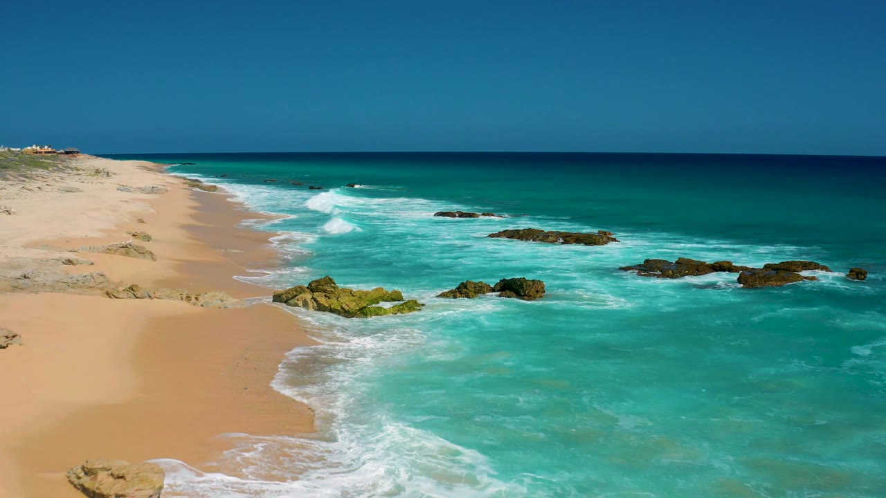 A later view of the El Cardoncito coast shows white waves wrapping around dark rocks, open sand at left, deep blue water near the horizon, and no visible people.