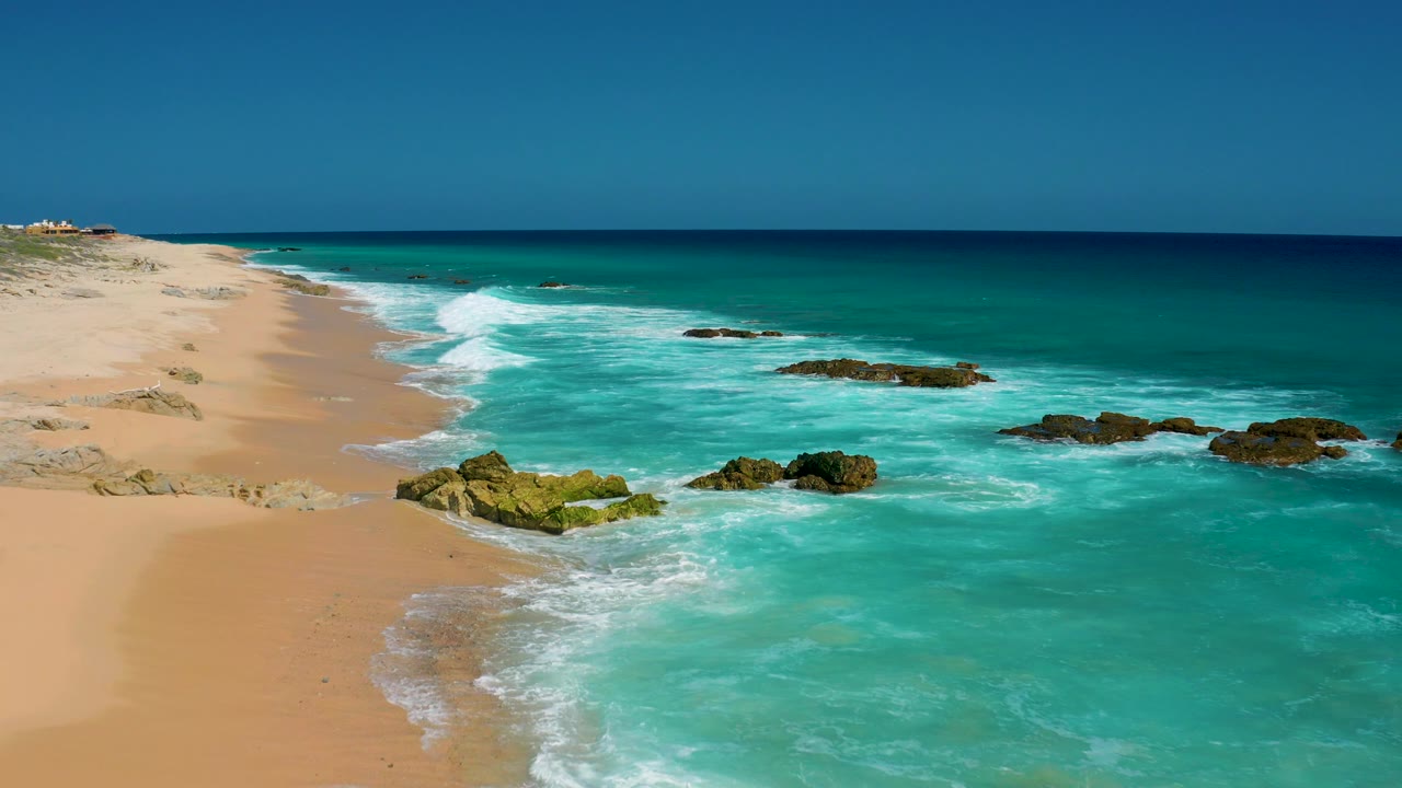 The El Cardoncito beach appears from a slightly shifted aerial composition, with more open water at right, rocks across the surf line, sand at left, and no visible people.