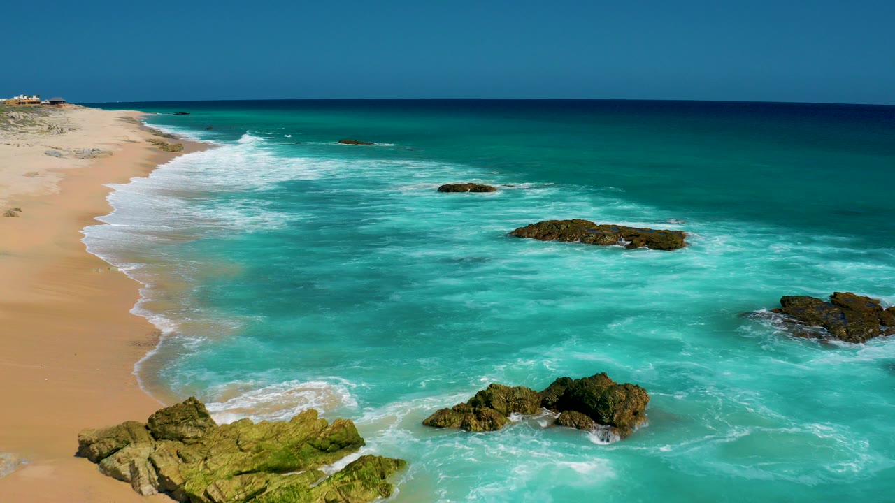 Teal ocean waves breaking around rocks beside a sandy beach.
