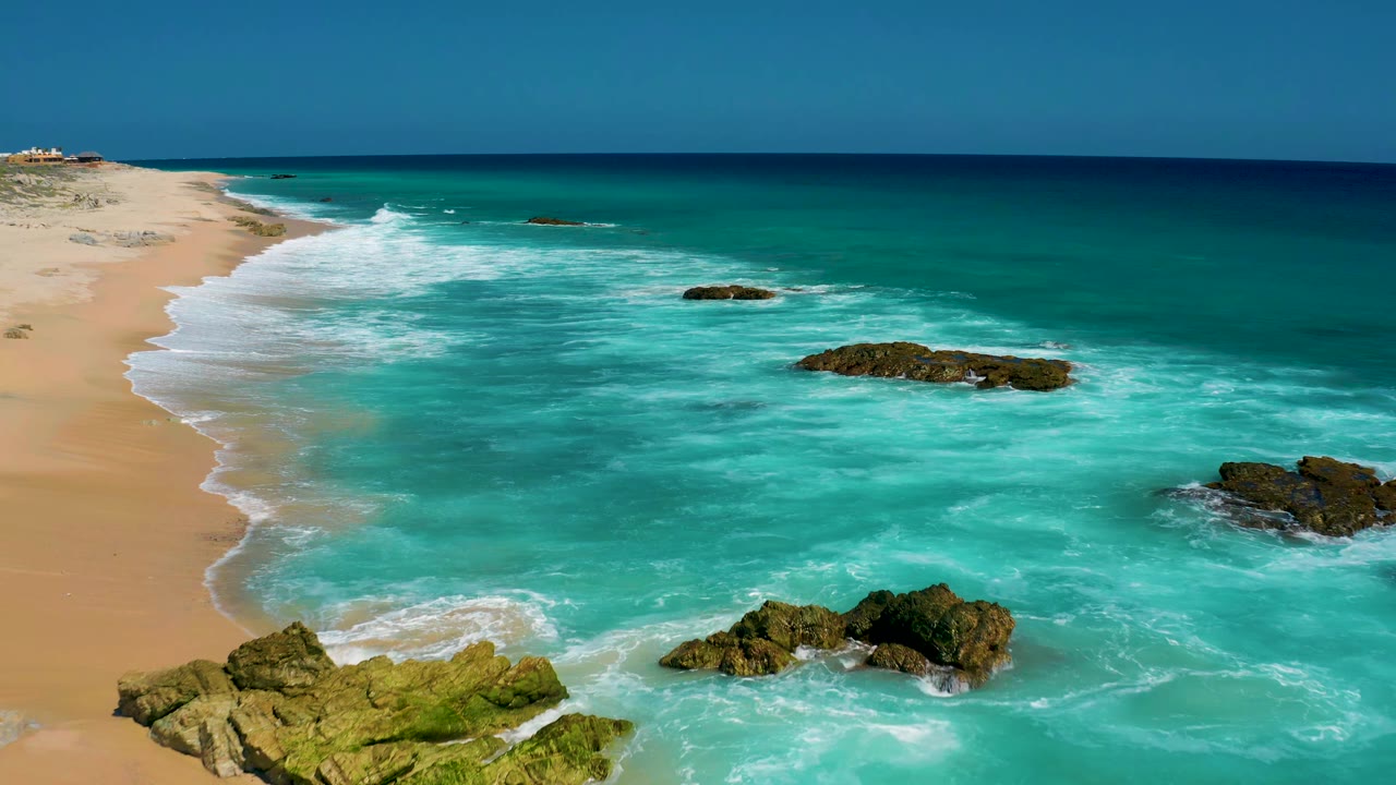 The drone-height view holds the sandy El Cardoncito shoreline on the left while pale surf spreads across shallow turquoise water, with no visible people.