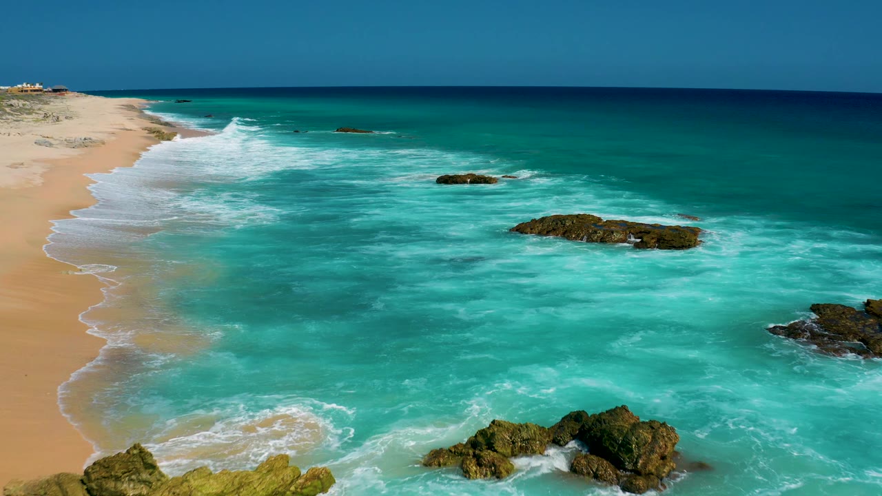 Turquoise water fills more of the frame as waves wrap around several dark rock outcrops off the El Cardoncito beach, with no visible people.