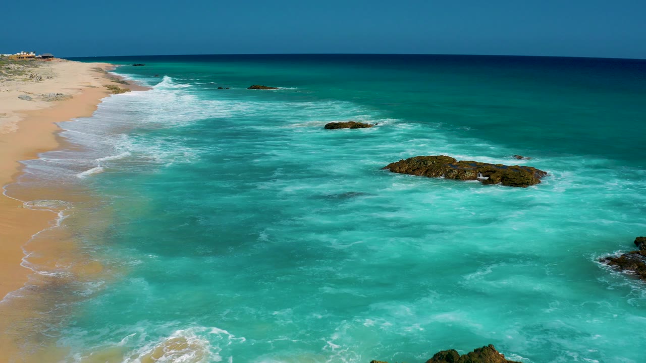 The left edge shows empty sand and a receding shoreline while teal waves move across scattered rocks in the water at El Cardoncito, with no visible people.