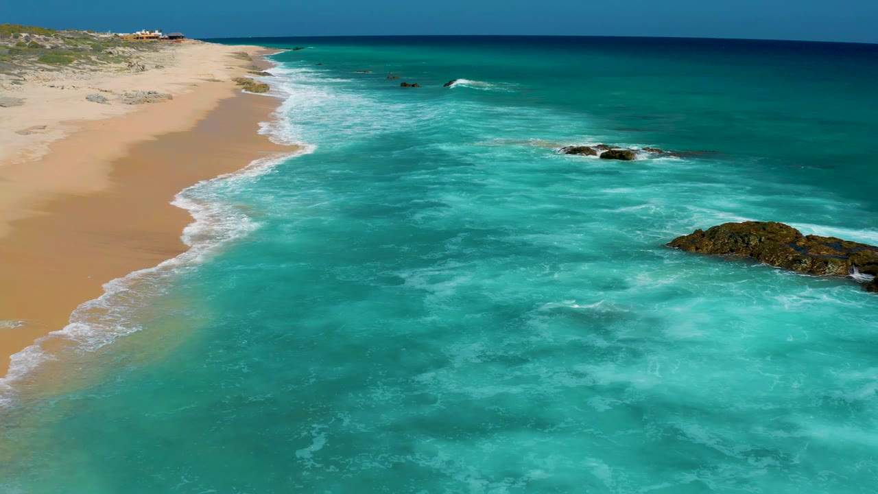 A high aerial frame over El Cardoncito shows the shoreline curving into the distance, pale sand at left, clear turquoise water across the frame, offshore rocks, and no visible people.