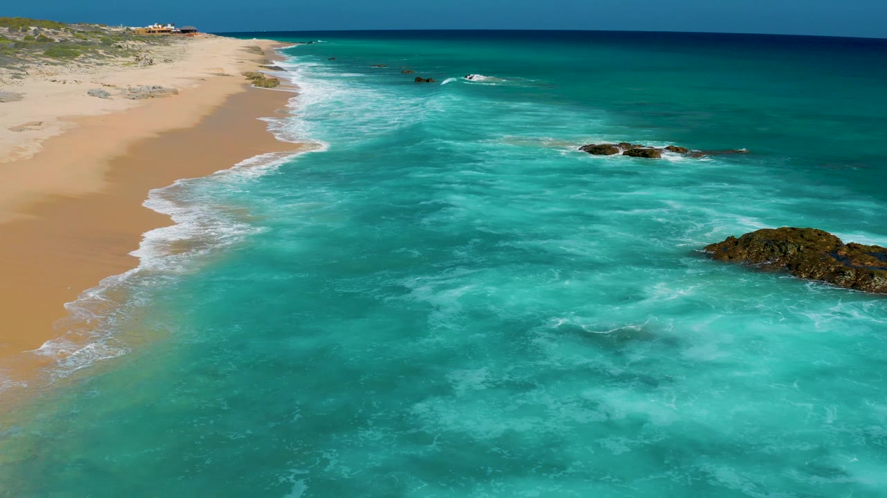 Aerial view over El Cardoncito with more open turquoise water filling the foreground, sandy beach along the left edge, rocks breaking the surf to the right, and no visible people.