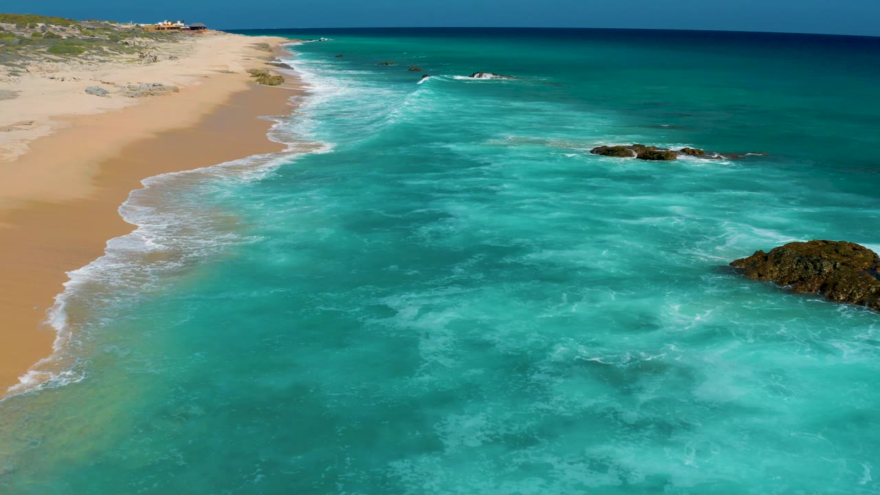 Elevated drone view at El Cardoncito with the sandy beach running diagonally at left, milky turquoise surf in the foreground, a rock cluster at right, and no visible people.