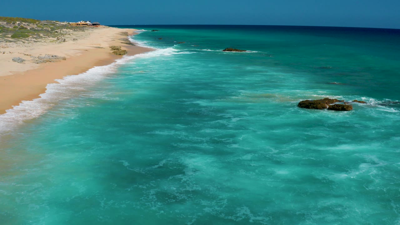 Aerial frame over El Cardoncito showing turquoise shallows in the foreground, a sandy beach on the left, white surf along the shore, dark rocks offshore, and no visible people.
