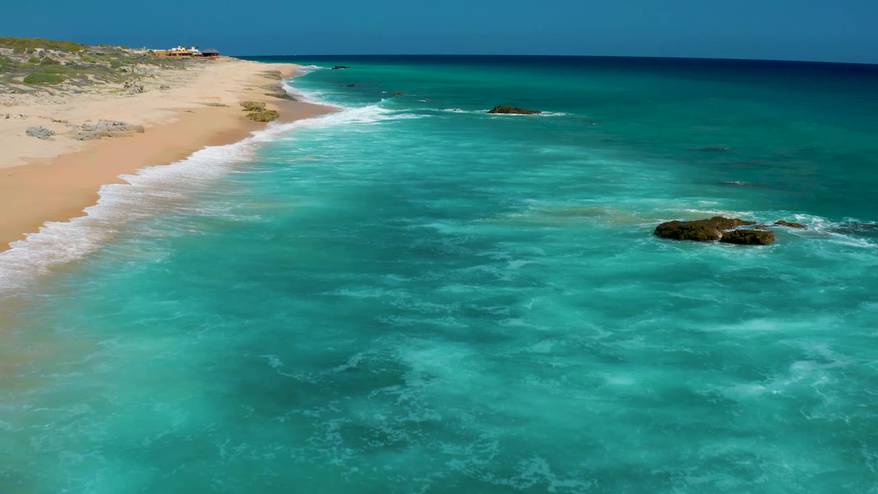 Elevated coastline view with clear blue-green water, pale sand, surf, and rocks offshore.