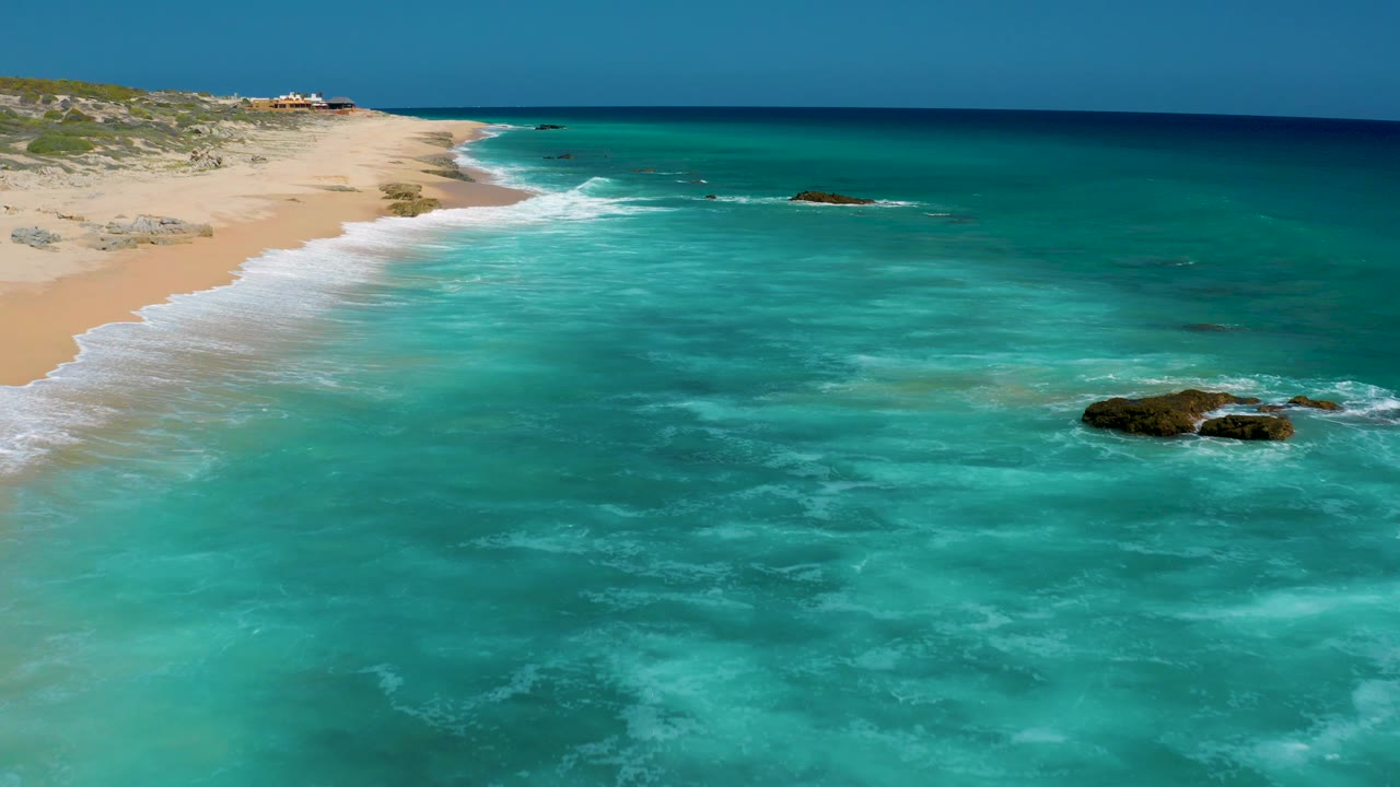 Aerial view of El Cardoncito with the sandy beach narrowing into the distance, turquoise water filling most of the frame, a dark rock cluster near the right edge, and no visible people.