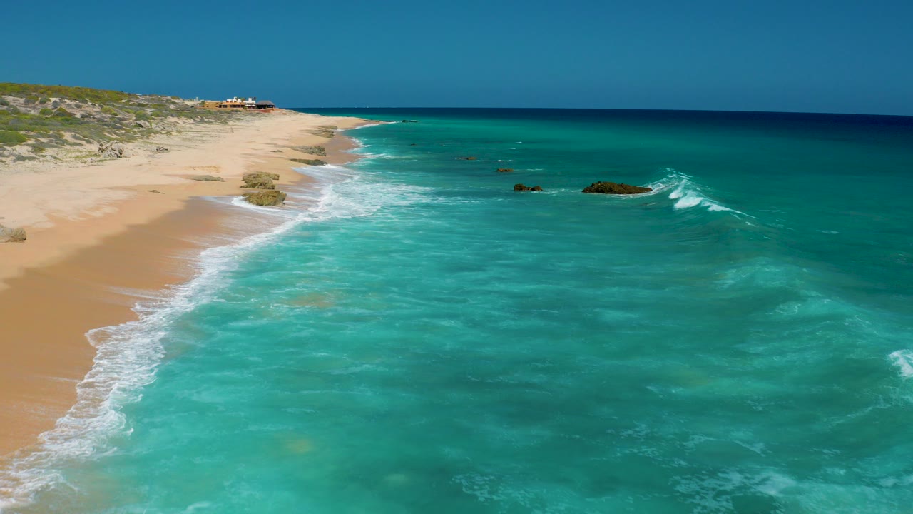Aerial frame from El Cardoncito, Baja California Sur, showing turquoise water washing onto a long sandy beach with low dunes inland and no visible people.