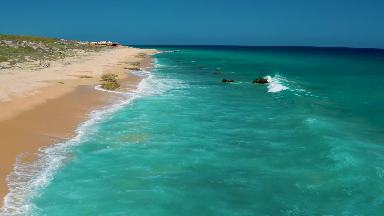 El Cardoncito aerial frame with the shoreline stretching into the distance, pale sand at left, open turquoise water at right, scattered rocks, and no visible people.