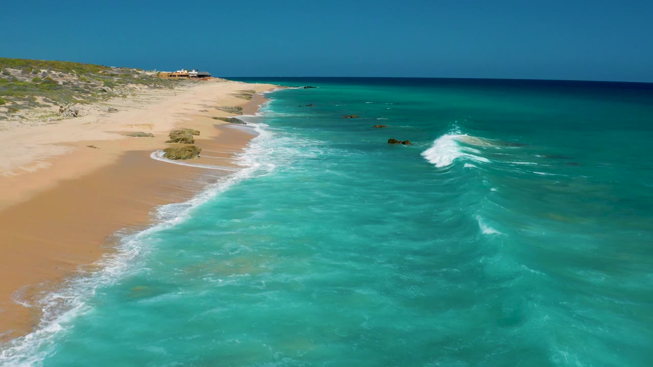 Aerial frame from El Cardoncito with a white wave line forming in turquoise water, sand and low vegetation along the left edge, and no visible people.
