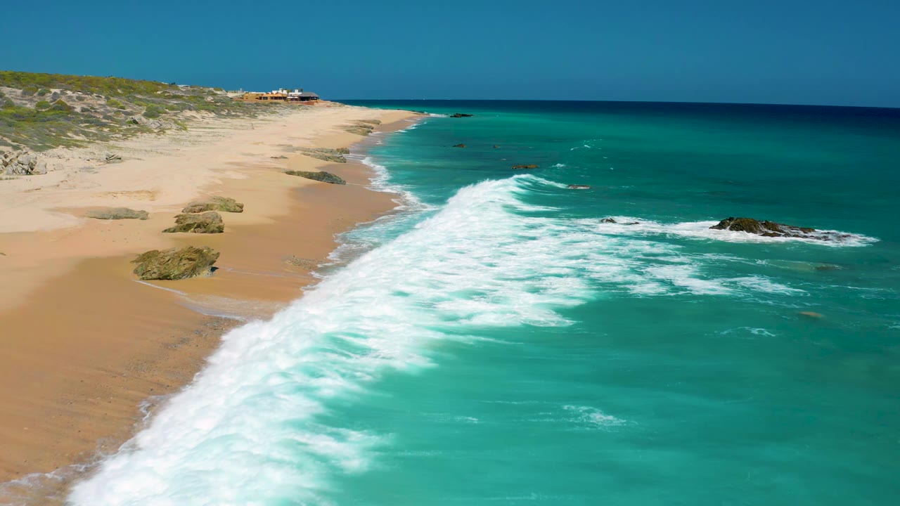 Aerial view in El Cardoncito showing a stronger band of whitewater crossing the lower frame, empty tan sand on the left, dark offshore rocks, and no visible people.