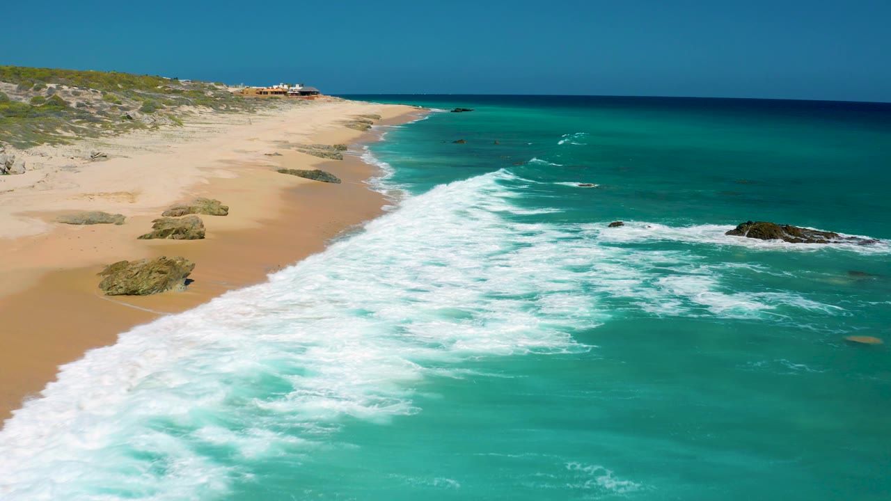 El Cardoncito aerial frame with broad white foam spreading over turquoise water, an empty sandy beach at left, rock outcrops near the shoreline, and no visible people.