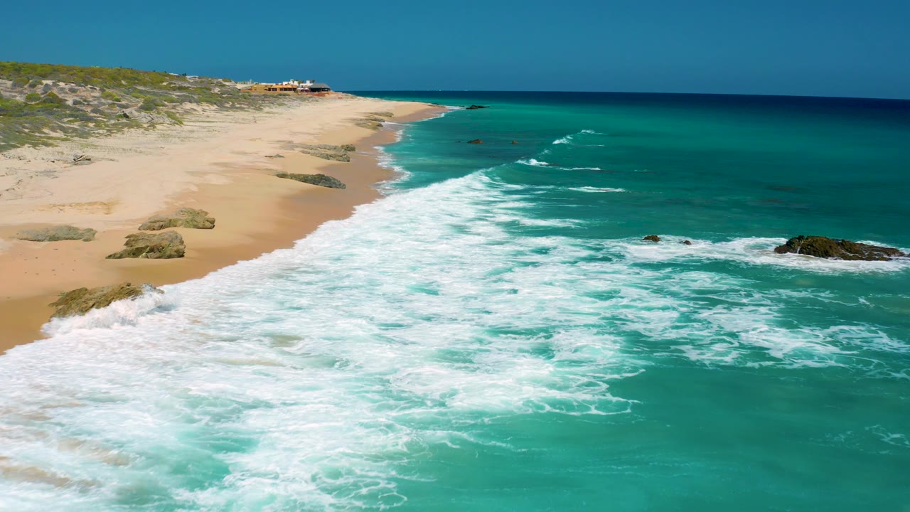 Aerial beach view in Baja California Sur with white surf filling the foreground, empty sand and scattered rocks to the left, blue-green water to the right, and no visible people.