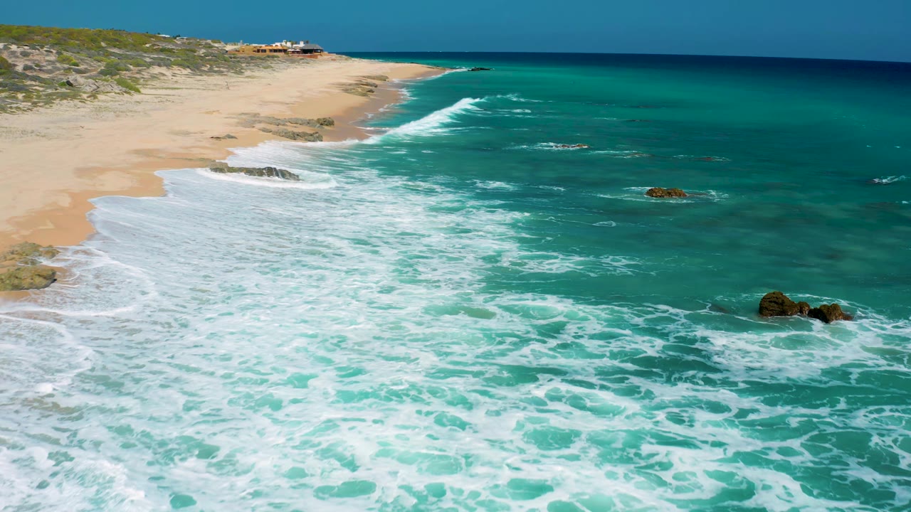 At El Cardoncito, Baja California Sur, white wave patterns widen across blue-green water while the pale beach and low dune vegetation remain at the left, with no visible people.