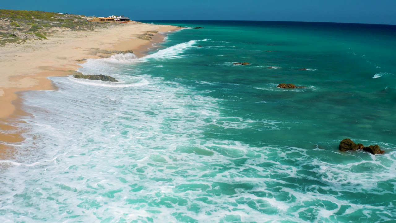 At El Cardoncito, Baja California Sur, a breaking wave spreads bright foam along the curved beach while scattered rocks sit in the turquoise shallows, with no visible people.