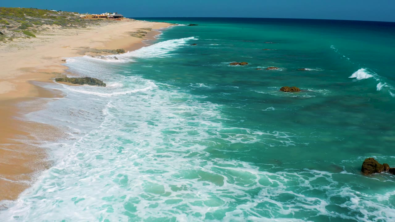 At El Cardoncito, Baja California Sur, the aerial composition looks farther along the beach as whitewater thins over tan sand and dark rocks break the surface, with no visible people.