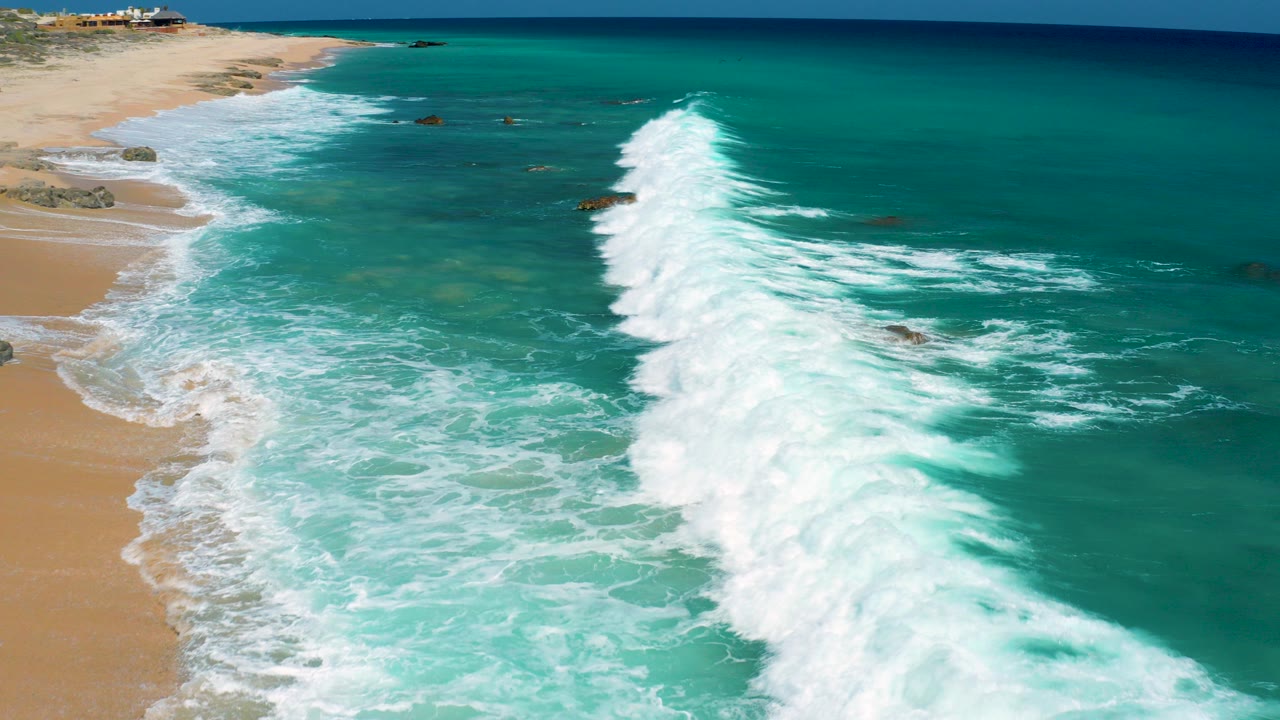 Aerial view over El Cardoncito, Baja California Sur, where white surf curves beside a sandy beach and turquoise water, with no visible people.