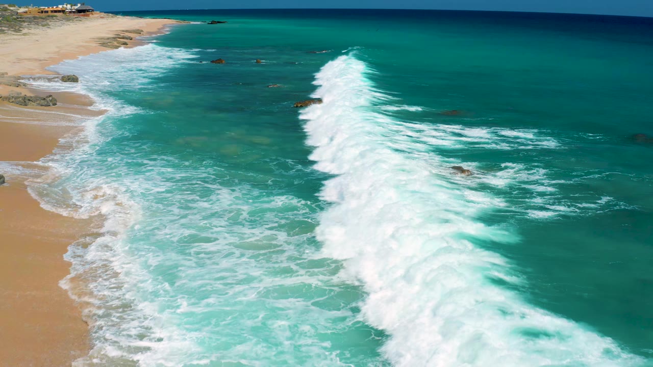 Aerial frame from El Cardoncito showing white breakers rolling across shallow turquoise water toward the sandy shoreline, with no visible people.