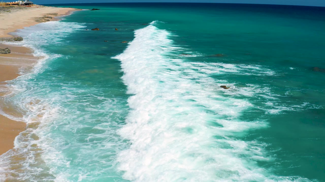 Aerial view at El Cardoncito with a larger white wave line spreading through blue-green water while the beach remains along the left edge, with no visible people.