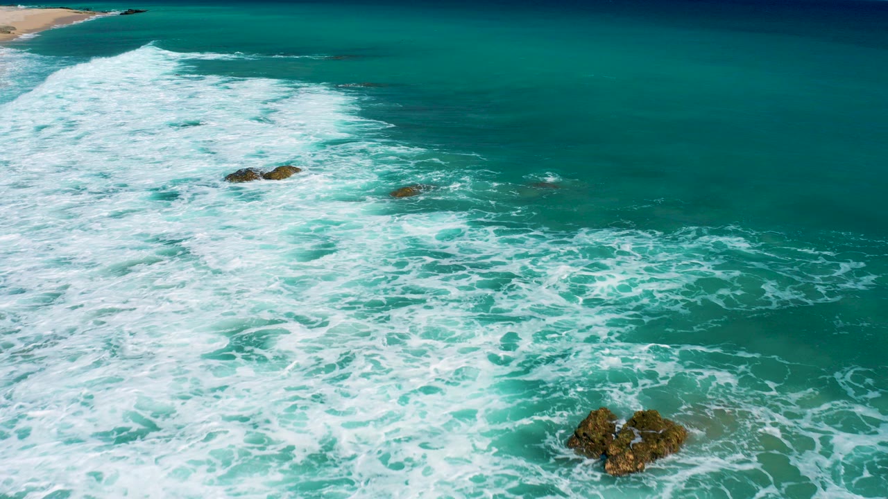 Aerial frame over El Cardoncito surf with white foam spreading across turquoise water, dark rocks in the lower and middle frame, a sandy beach edge at upper left, and no visible people.