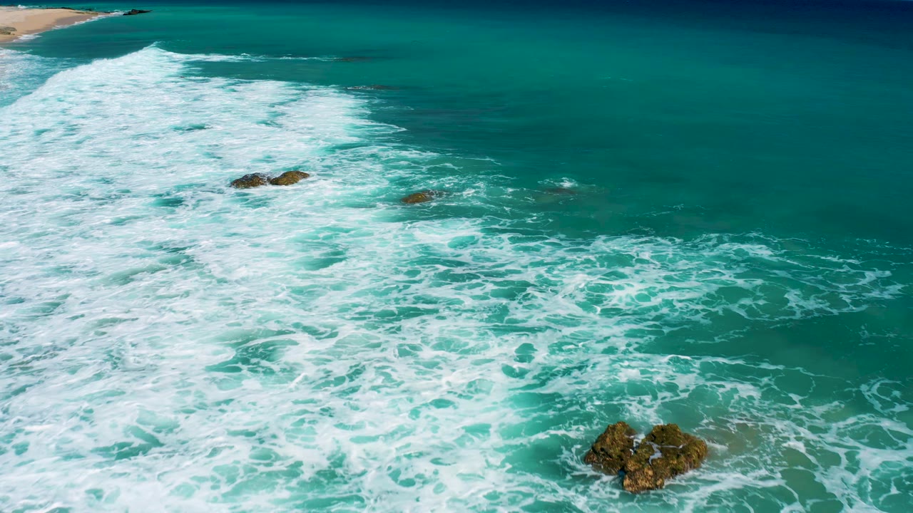 Aerial frame over El Cardoncito with foamy surf covering more of the foreground, dark rocks breaking the water surface near center and lower right, beach at upper left, and no visible people.