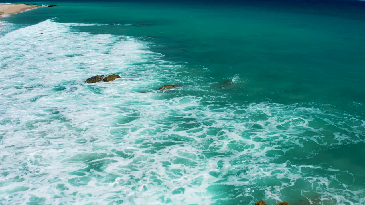 Aerial frame over El Cardoncito where white surf pushes diagonally from the beach side into turquoise water, with dark rocks near the center and lower edge and no visible people.