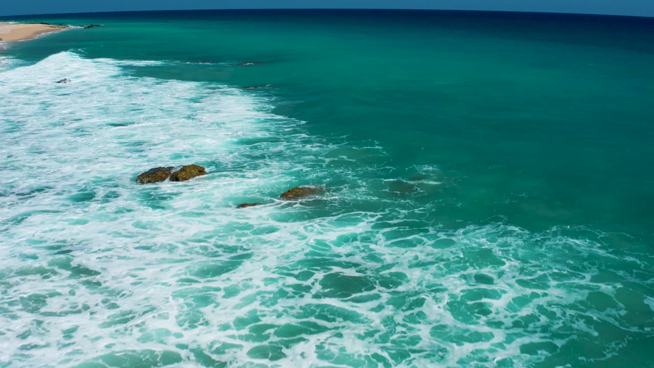 Aerial frame over El Cardoncito with the surf band running down the left side, darker rocks near center, clear turquoise water to the right, beach at upper left, and no visible people.