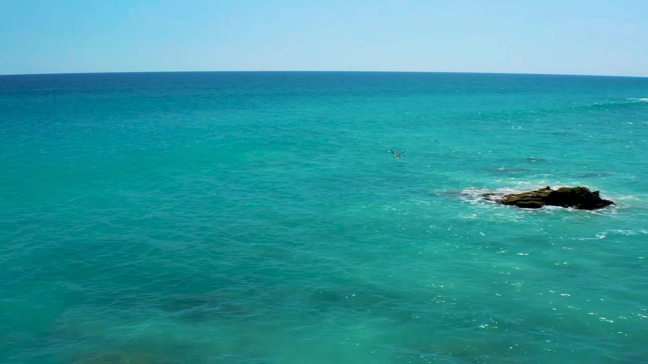 Blue-green ocean surface at El Cardoncito with a low dark rock surrounded by white foam, deeper blue water beyond, clear sky above, and no visible people.