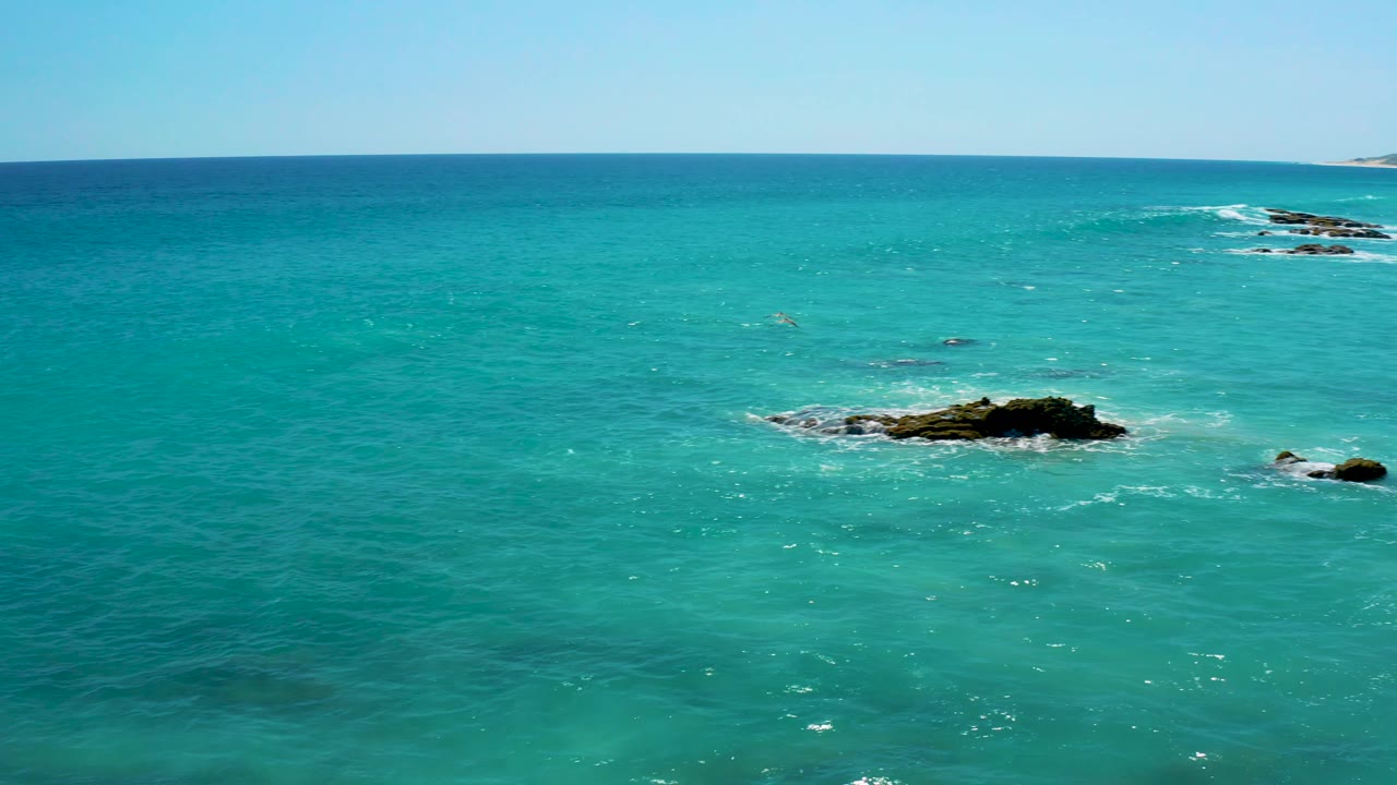 Clear turquoise water in Baja California Sur with a dark rock patch to the right, pale foam around the rocks, distant low shoreline at the far edge, and no visible people.