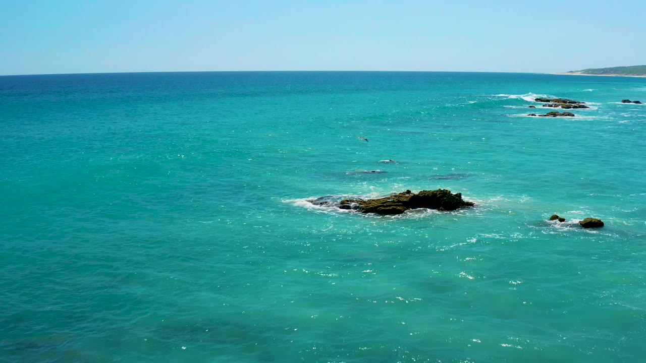 Wide elevated view over El Cardoncito water with scattered rocks, white foam streaks, shallow green tones, a low distant shoreline, and no visible people.