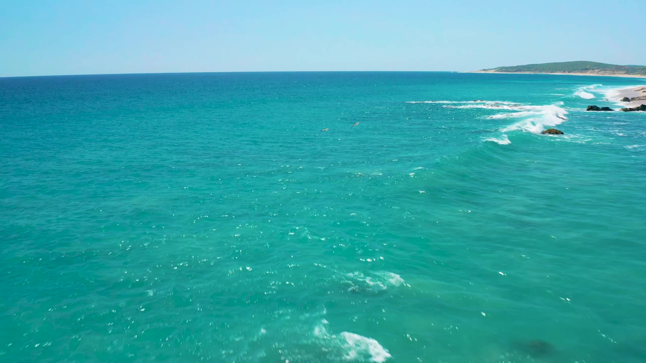 Elevated ocean view near El Cardoncito showing more open turquoise water in the foreground, small white breakers near the right edge, and no visible people.