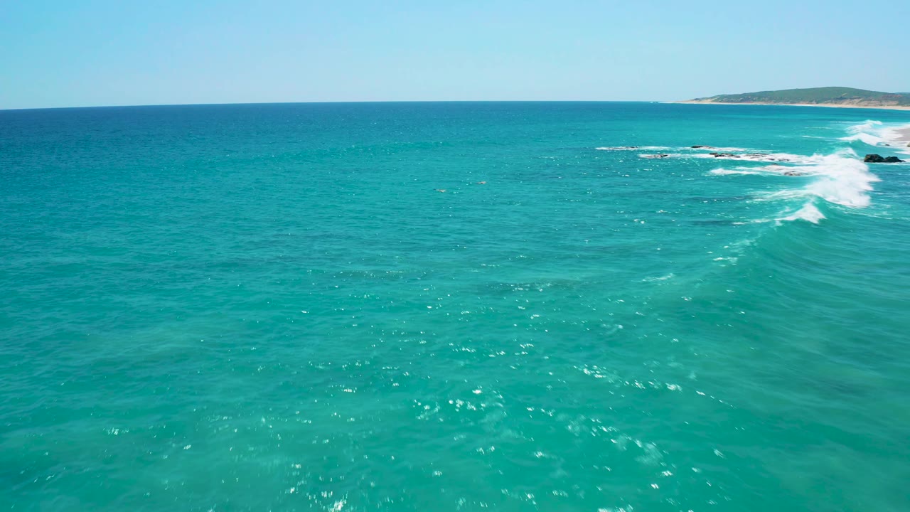 Aerial frame over bright blue-green sea near El Cardoncito, with a compact rocky surf line at the far right and no visible people.