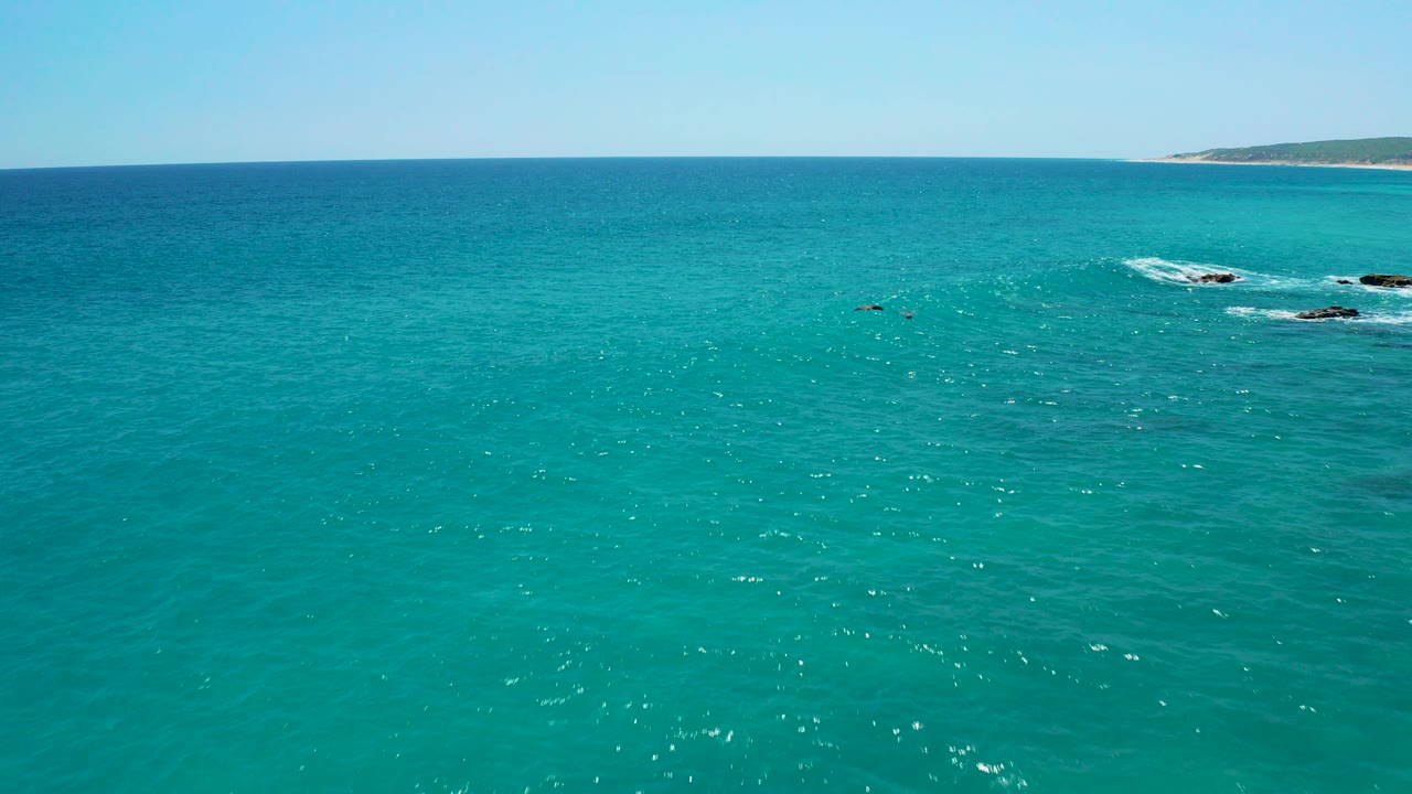 Aerial view over turquoise sea at El Cardoncito, Baja California Sur, with small white wave breaks near dark rocks, distant coastline on the horizon, and no visible people.