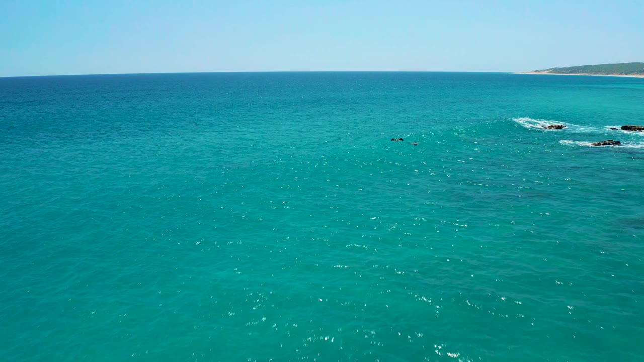 Aerial frame over blue-green water at El Cardoncito, Baja California Sur, with foamy surf around dark rocks slightly right of center and no visible people.
