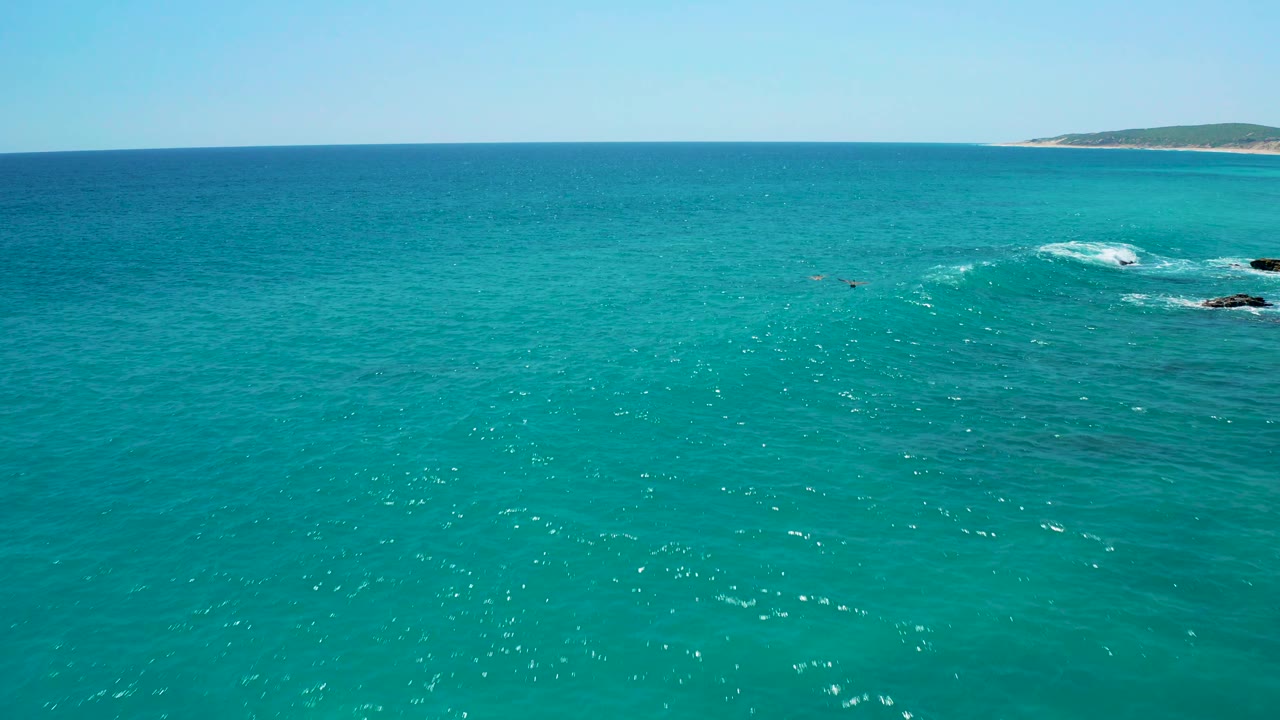 Wide aerial view at El Cardoncito, Baja California Sur, showing turquoise ocean, a white surf patch over rocks at the right edge, low coastline, and no visible people.