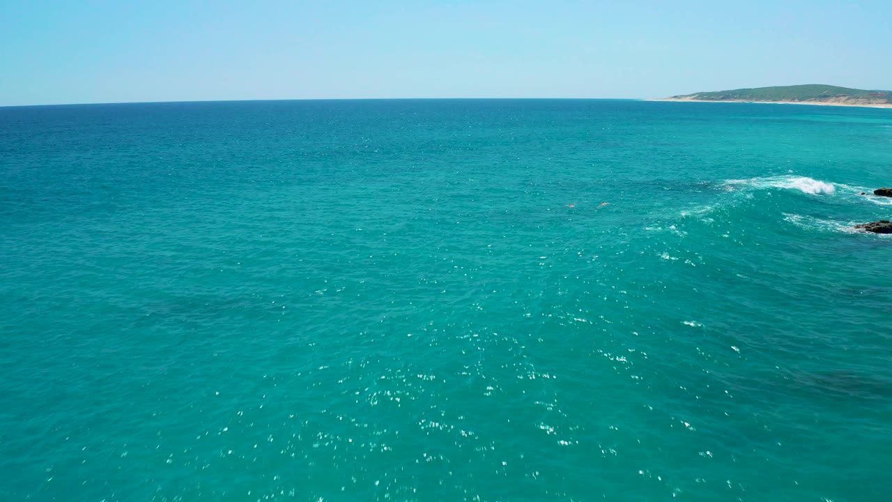 Aerial sea view from El Cardoncito, Baja California Sur, with a white wave line crossing dark rocks, sandy shoreline in the far background, and no visible people.