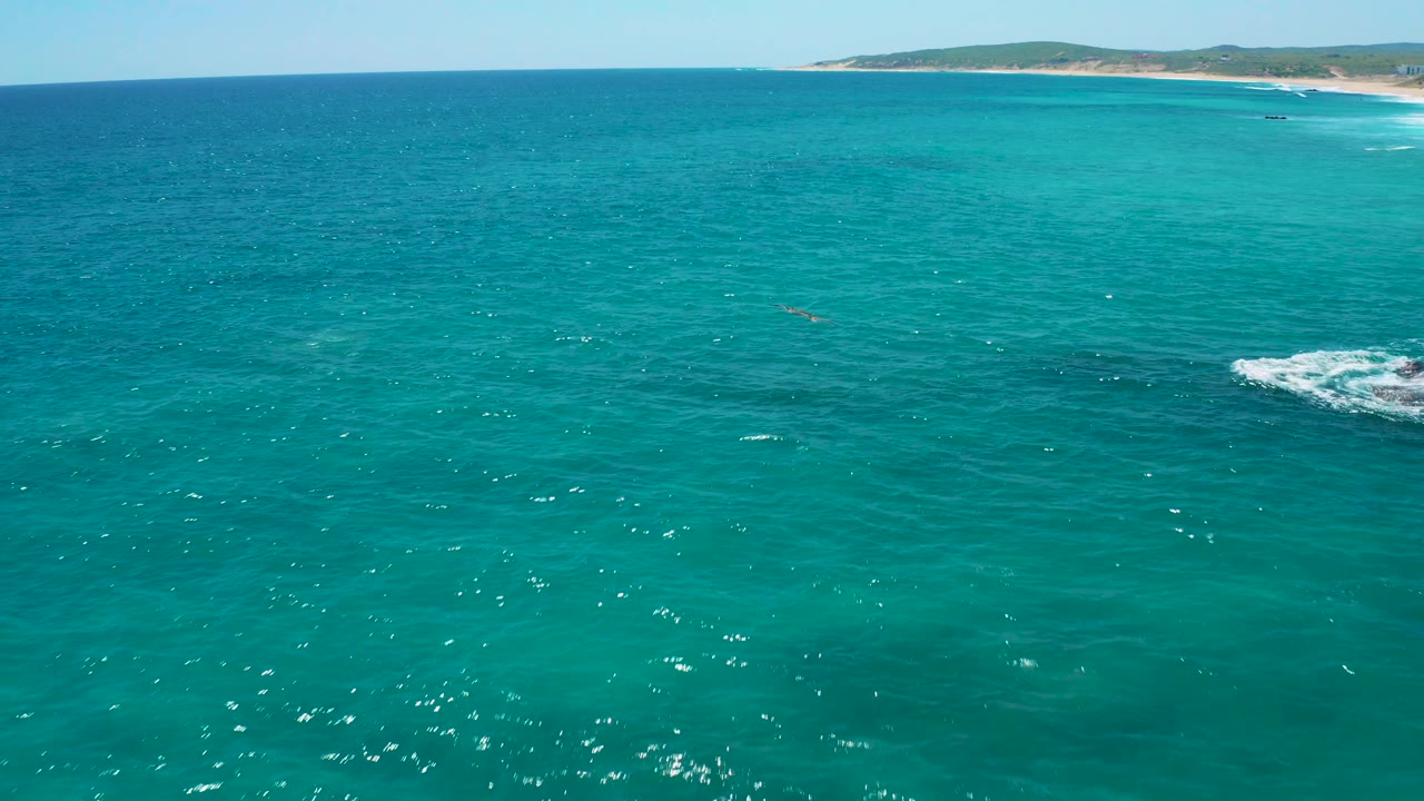 Wide aerial view at El Cardoncito showing turquoise water, a sandy shoreline across the upper right, distant low hills, and no visible people.