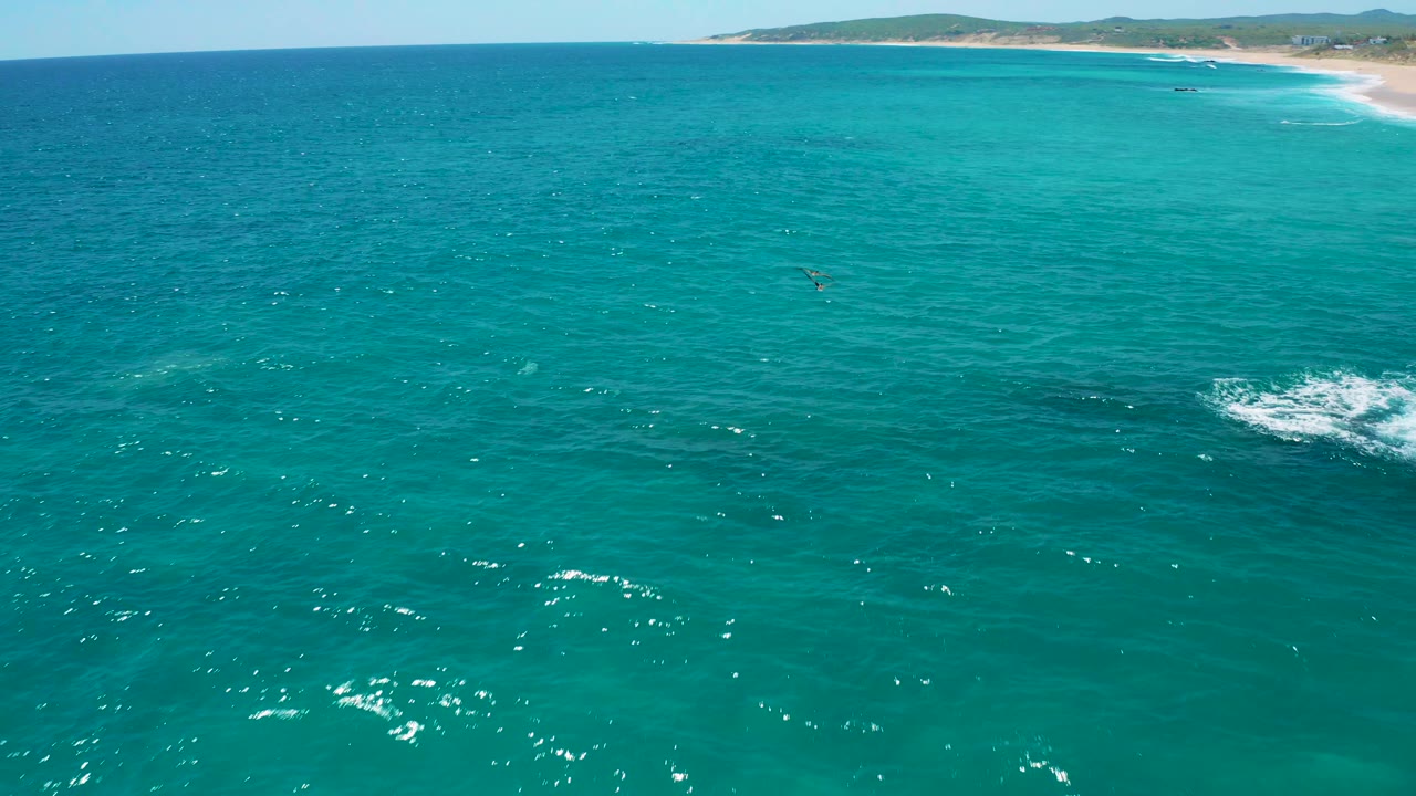 Aerial view at El Cardoncito with clear turquoise water in front, a sandy beach on the horizon, white water near the lower right, and no visible people.