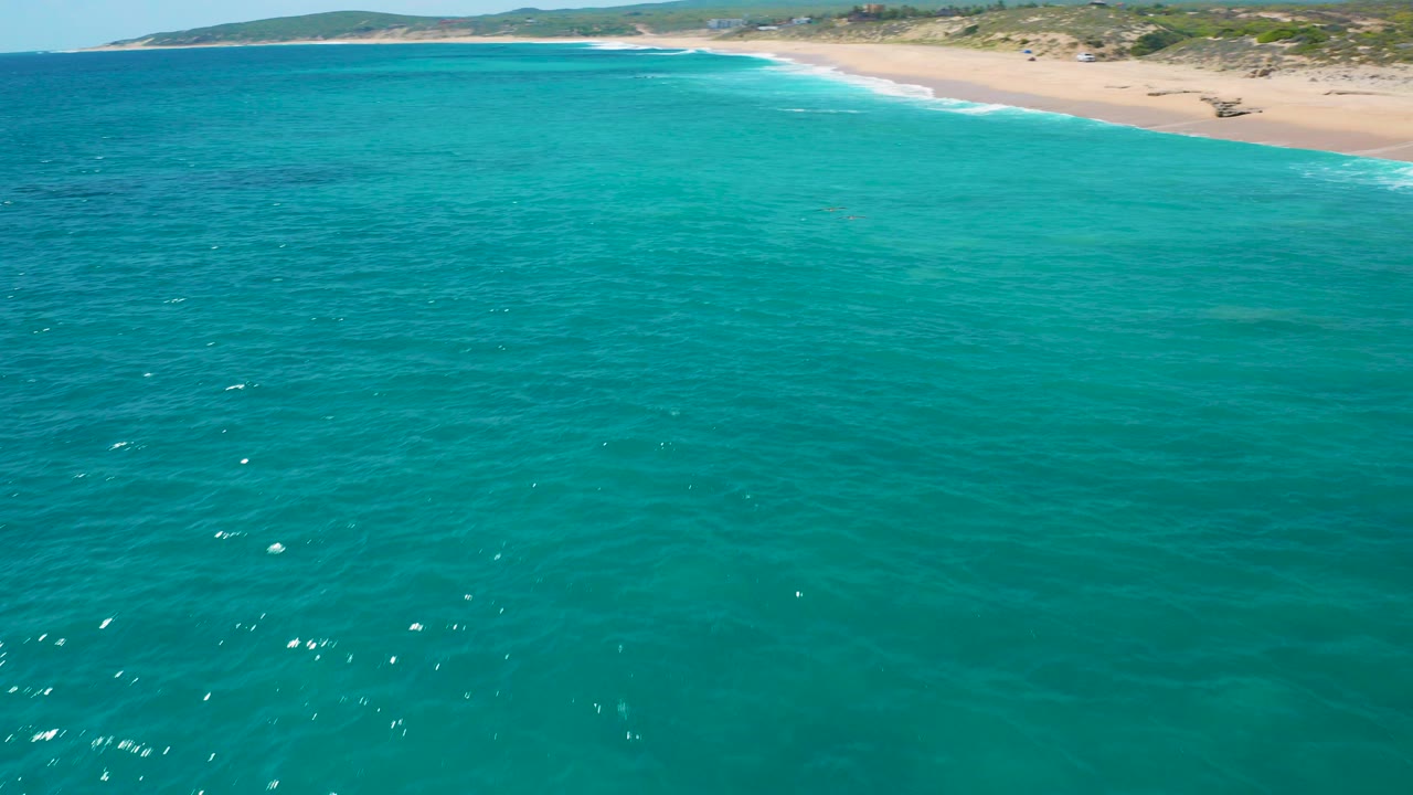 Aerial view over turquoise water toward a sandy beach, low green dunes, and distant hills in El Cardoncito, Baja California Sur, with no visible people.
