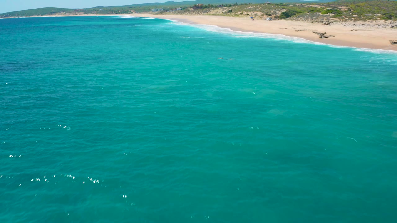 Clear blue-green water fills the foreground, with pale sand, small breaking waves, low dunes, and distant hills in El Cardoncito, Baja California Sur, with no visible people.
