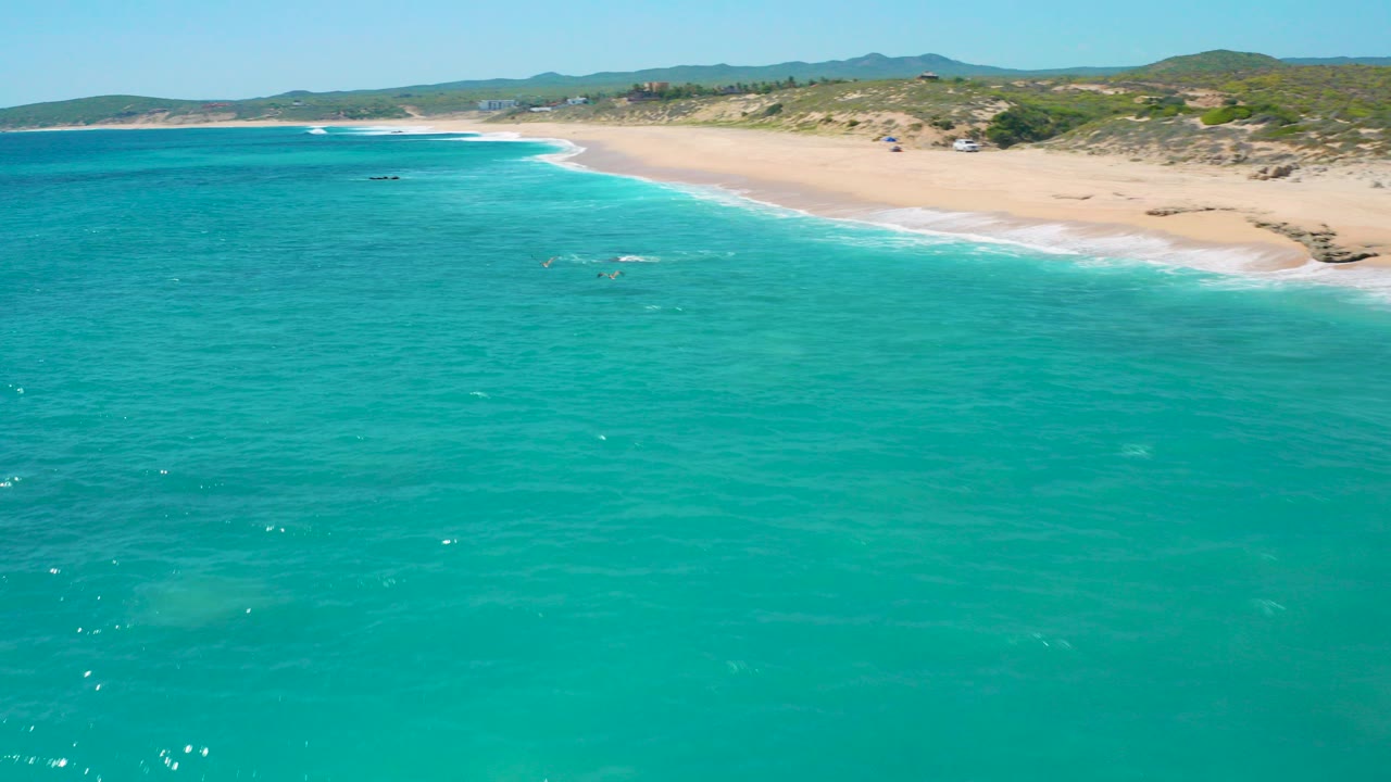Aerial view at El Cardoncito, Baja California Sur, with turquoise water filling the foreground, a pale beach curving across the upper frame, low dunes and hills behind it, and no visible people.