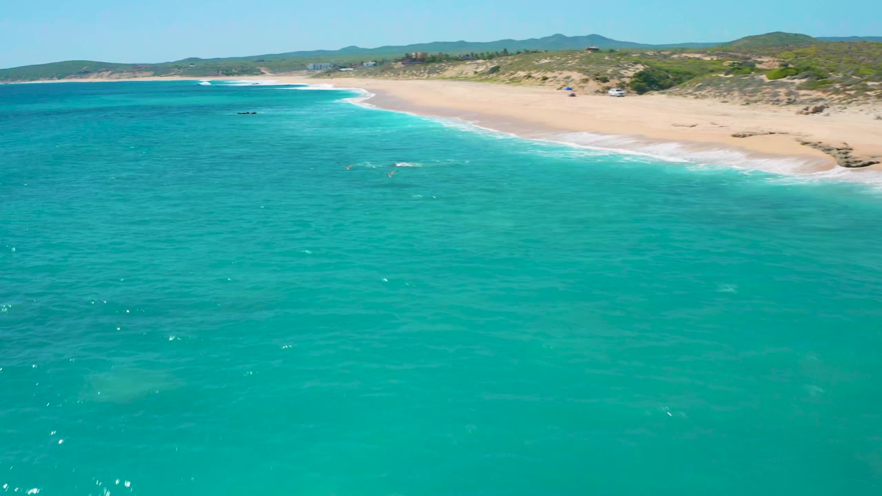 Aerial frame over blue-green water at El Cardoncito, showing the beach running along the upper right, a thin white surf edge, low vegetation, distant hills, and no visible people.