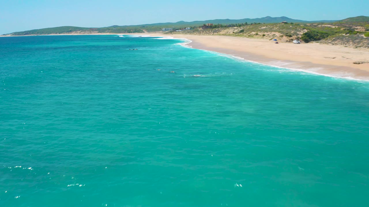 High offshore view at El Cardoncito with broad turquoise water in front, a long sand beach across the top, scattered low vegetation, distant hills, and no visible people.